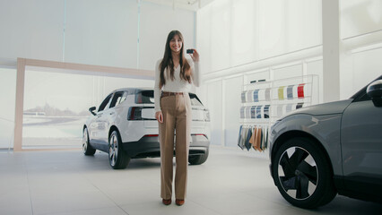 Caucasian woman standing in modern car showroom with large panoramic windows. Holding car keys in one hand. White electric car and part of gray vehicle visible in camera. Dealership concept.