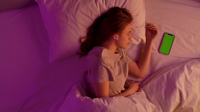 Young woman sleeping in bed, laying head on the pillow covered with blanket in pink neon light, wearing pods, smartphone with chroma key green screen background on the bed. Top view.