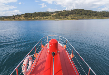 View From the Bow of a Red Boat on Calm Waters, With a Forested Hillside in the Background Under a Partly Cloudy Sky