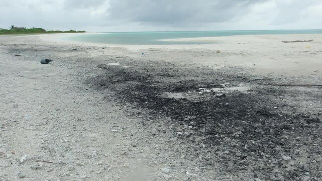 A patch of burnt debris scars the white sand of a Maldives sandbank. The turquoise ocean and overcast sky contrast
