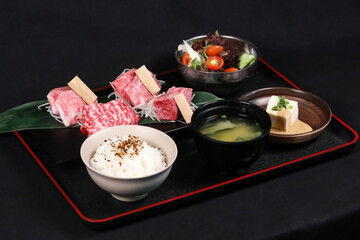 Wagyu Beef Yakiniku Bento Set on a black tray include raw Wagyu beef slices, rice, miso soup, tofu, and salad isolated on dark background side view of Japanese cuisine