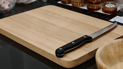 A close-up shot captures a wooden cutting board with a knife resting on top, set against a dark kitchen countertop. A wooden bowl is partially visible in the foreground and clear food containers © dongwei