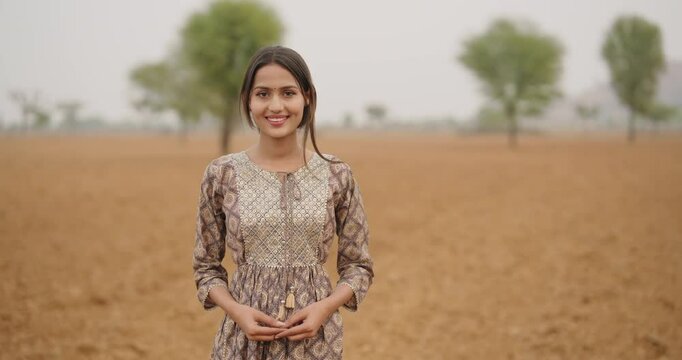 Indian young adult gen z farmer woman lady wear casual dress outdoor farmland looking at camera asian happy smiling village teen age girl fold hand posing for cam outside ground