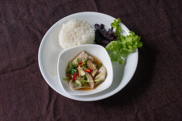Steamed Seabass Fish with Soy Sauce is served with rice and greens. The fish, with ginger and chili, sits in a soy-based sauce. Rice and lettuce are on a white plate against a brown fabric.