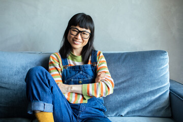 Portrait of a happy Asian woman wearing overalls sitting on couch and smiling at home