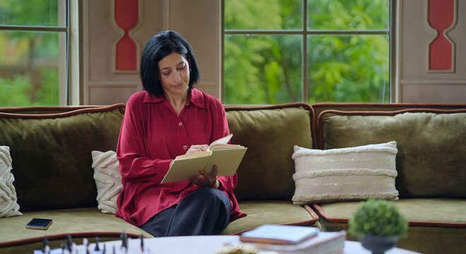 Indian happy relax gen x old aged woman lady sitting on cozy sofa couch hold book read good story indoor room place. Smiling mature adult elder female spend free time enjoy day inside living home - Powered by Adobe