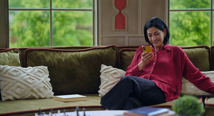 Indian happy relax gen x woman lady sitting sitting on cozy sofa couch hold mobile typing text sms...