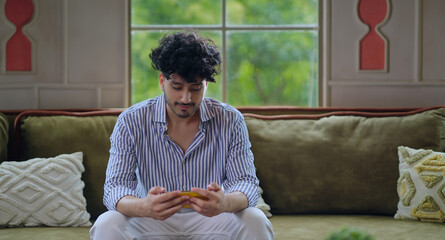 Indian happy young adult gen z man guy sitting on cozy sofa couch use mobile look screen play casual online video game win enjoy day. Teen age male hold phone have fun joy spend time at indoor house