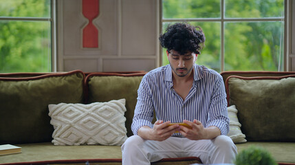 Indian upset young adult gen z gamer man guy sitting on cozy sofa couch use mobile look screen play...