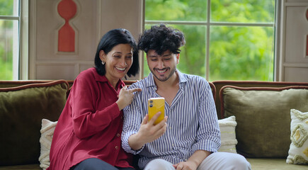 Indian Asian happy older woman mom lady young adult man male son sitting cozy sofa couch use mobile phone talk video call make funny gossip look cam screen joy enjoy fun day spend time indoor house