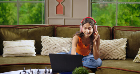 Indian happy young adult gen z women lady sitting cozy sofa couch sing song use laptop switch...