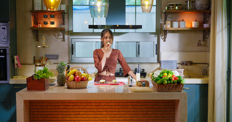 Indian happy young adult gen z smiling woman lady standing indoor modern kitchen hungry teen age girl female eat fresh apple fruit diet food spend time enjoy day