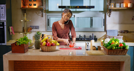 Indian young adult gen z woman lady use mobile talk phone call distant enjoy day time have fun joy teen girl hold knife phone speak gossip cut fresh tomato slice on chop board indoor modern kitchen