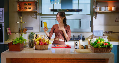 Indian happy young adult woman lady hold mobile talk on video call indoor home teen age girl female use phone look screen enjoy distant gossip speak funny spend day time have fun joy kitchen counter