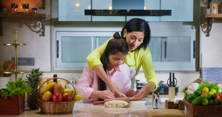 Indian happy woman lady with cute teen age cute girl rolling raw flour dough learn cooking food indoor home smiling young mom teach to kid child baking cookies enjoy day indoor modern home kitchen