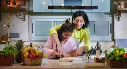Indian happy woman lady with cute teen age cute girl rolling raw flour dough learn cooking food indoor home smiling young mom teach to kid child baking cookies enjoy day indoor modern home kitchen