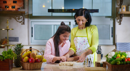 Indian happy woman lady with cute teen age cute girl rolling raw flour dough learn cooking food indoor home smiling young mom teach to kid child baking cookies enjoy day indoor modern home kitchen