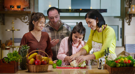 Indian mom chef wife hold knife teach to teen age girl child cut fresh vegan slice on chop board indoor modern home kitchen happy dad man with young female talk enjoy learn have fun joy spend day time