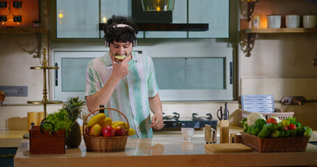 Indian happy young adult gen z man guy listen online mp3 song music have fun joy indoor kitchen teen boy male hold knife cut slice piece of fresh apple eat healthy diet enjoy spend day time