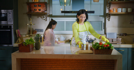 Indian mom standing wear apron and hand gloves collect pick up waste trash in box day time adult woman lady do work teen age girl child use rag wipe table dust keep fresh neat tidy clean kitchen home