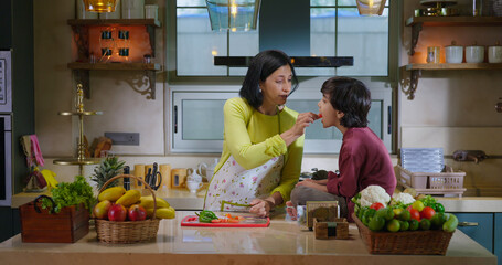 Indian happy mature mom standing indoor modern home kitchen hold knife cut fresh slice on chop board young adult woman lady talk gossip son child boy eat vegan piece enjoy day spend time have fun joy