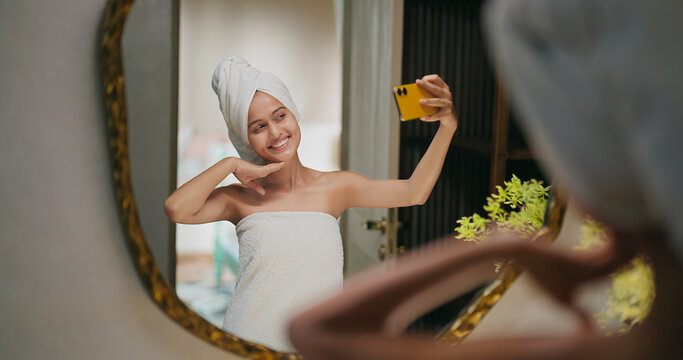 Indian happy gen z young adult female lady wrapped towel head and body after shower use mobile talk gossip video call smiling teen girl hold phone look cam mirror take selfie enjoy fun joy day house