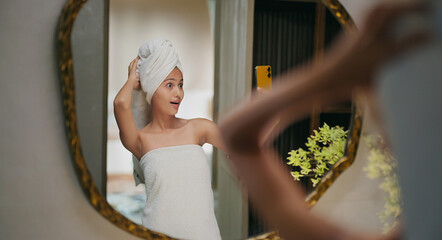 Indian happy gen z young adult female lady wrapped towel head and body after shower use mobile talk gossip video call smiling teen girl hold phone look cam mirror take selfie enjoy fun joy day house