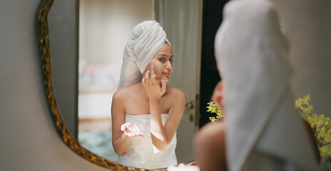 Indian young adult gen z woman lady wrapped towel on head and body look mirror apply make up cream on dry cheek face teen age girl female hold bottle take liquid hand do fresh day skin care house home