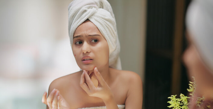 Indian upset young adult gen z woman lady wrapped towel on head and body look mirror feel sad facial pores skin dry cheek teen girl female worried for acne pimple touch face heath care at indoor home