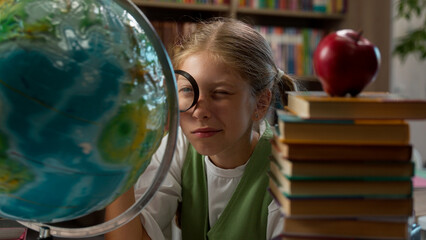 Curious funny adorable intelligent Caucasian little girl playful schoolgirl child school kid pupil learner student looking globe through magnifying glass studying education world map exploring indoor