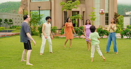 Indian happy family young man woman group play ball game throwing in air have fun joy enjoy outdoor park garden Playful cute teen girl son child kid boy with adult mom lady dad spend day time outside	
