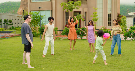 Indian happy family young man woman group play ball game throwing in air have fun joy enjoy outdoor park garden Playful cute teen girl son child kid boy with adult mom lady dad spend day time outside