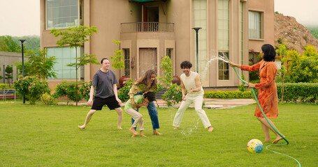 Indian happy mom hold hose pipe spray water on young adult family teen girl dad cute son kid boy child male man female group play ball game have fun joy spend time enjoy day trip house lawn park