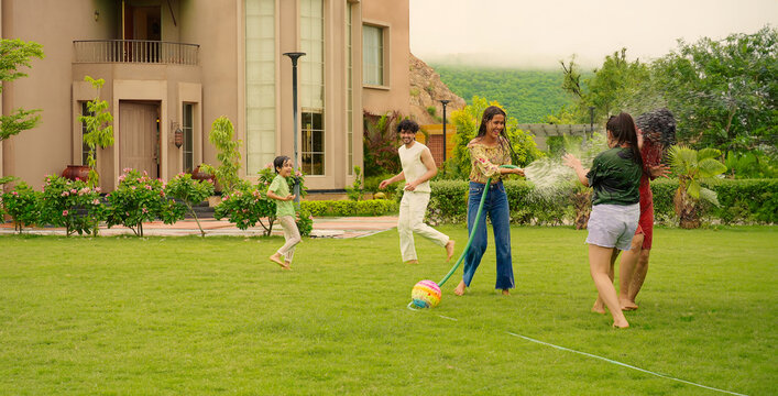 Indian happy teen girl lady mom hold hose pipe spray water on family group play game house lawn cute kid boy son child young adult man have fun joy day spend funny time enjoy trip outside picnic park