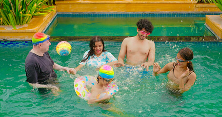 Indian wet family teen age child group standing in pool playing splash water have fun joy spend day time water park happy father dad young adult girl kid boy wear ring enjoy hotel trip resort picnic