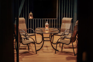 A cozy outdoor patio at night with a glass-top table and four chairs. A lantern with a candle casts a warm glow, creating a relaxing ambiance on the wooden deck.