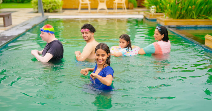 Indian happy wet family male female lady man group do warm up exercise in pool enjoy day outdoor water park resort trip picnic young mom dad with cute teen girl child adult boy have fun joy spend time