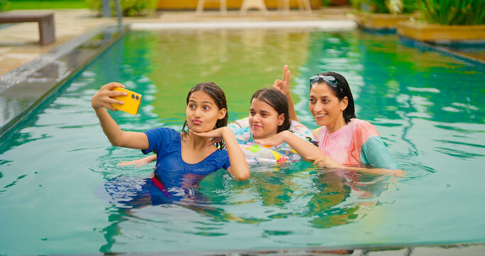 Indian happy wet cute teen girl young mom hold mobile look cam take selfie do funny pose in water pool resort adult mother child use phone record shoot video spend time have fun joy outdoor hotel trip