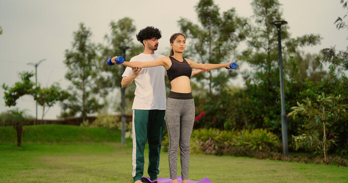 Indian young adult man woman use dumbbells do hand lifting muscle workout outdoor garden park healthy fit boy male guy standing teach to female girl good habit learn gym warm up self health body care
