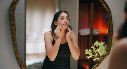 Indian gen x senior mature adult older elder short hair woman lady look mirror use beauty apply soft cream on dry face female mom hold liquid in hand do fresh day skin care at house home