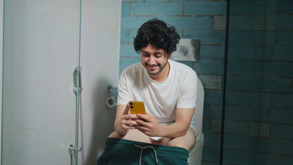 Indian Asian active young adult gen z happy man guy sitting on toilet use mobile phone online share...