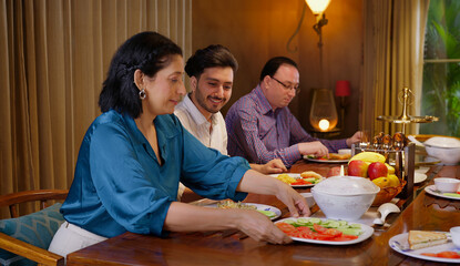 Indian family group sitting around dining table chair enjoy night dinner indoor home happy mom lady serve fresh salad cute kid boy dad hold spoon eat tasty food talk gossip have fun joy spend time
