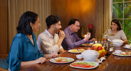 Indian family group sitting around dining table chair enjoy night dinner indoor home happy mom lady serve fresh salad cute kid boy dad hold spoon eat tasty food talk gossip have fun joy spend time