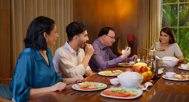 Indian family group sitting around dining table chair enjoy night dinner indoor home happy mom lady serve fresh salad cute kid boy dad hold spoon eat tasty food talk gossip have fun joy spend time