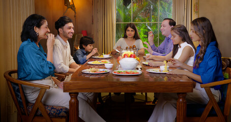Indian happy family group with cute kid child sitting around dining table chair enjoy night dinner adult dad teen girl young boy talk mom lady serve fresh salad eat tasty food have fun joy spend time