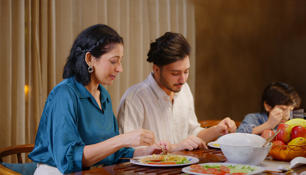 Indian family group sitting around dining table chair enjoy night dinner indoor home. happy mom lady feed with hand serve fresh salad cute kid boy hold spoon eat tasty food have fun joy spend time - Powered by Adobe