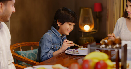 Indian happy young gen z woman lady with cute kid child sitting around dining table chair hold spoon eating tasty food adult female teen age male boy enjoy night dinner spend time indoor modern home