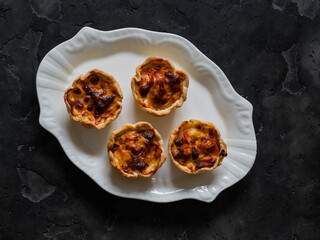 Mini pies with ham, dried tomatoes and cheese - a delicious snack, breakfast, appetizers on a dark background, top view