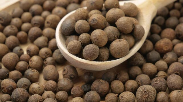 Grains of dried allspice in a wooden spoon close-up. Spices, black pepper peas on a wooden background