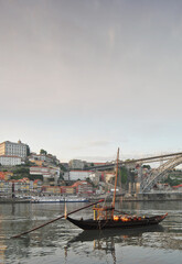 Obraz premium Portrait of downtown Porto in the late afternoon with a sunset sky, the Douro River with a traditional and old wine transport boat, part of the metal bridge of the Dom Luis metro bridge